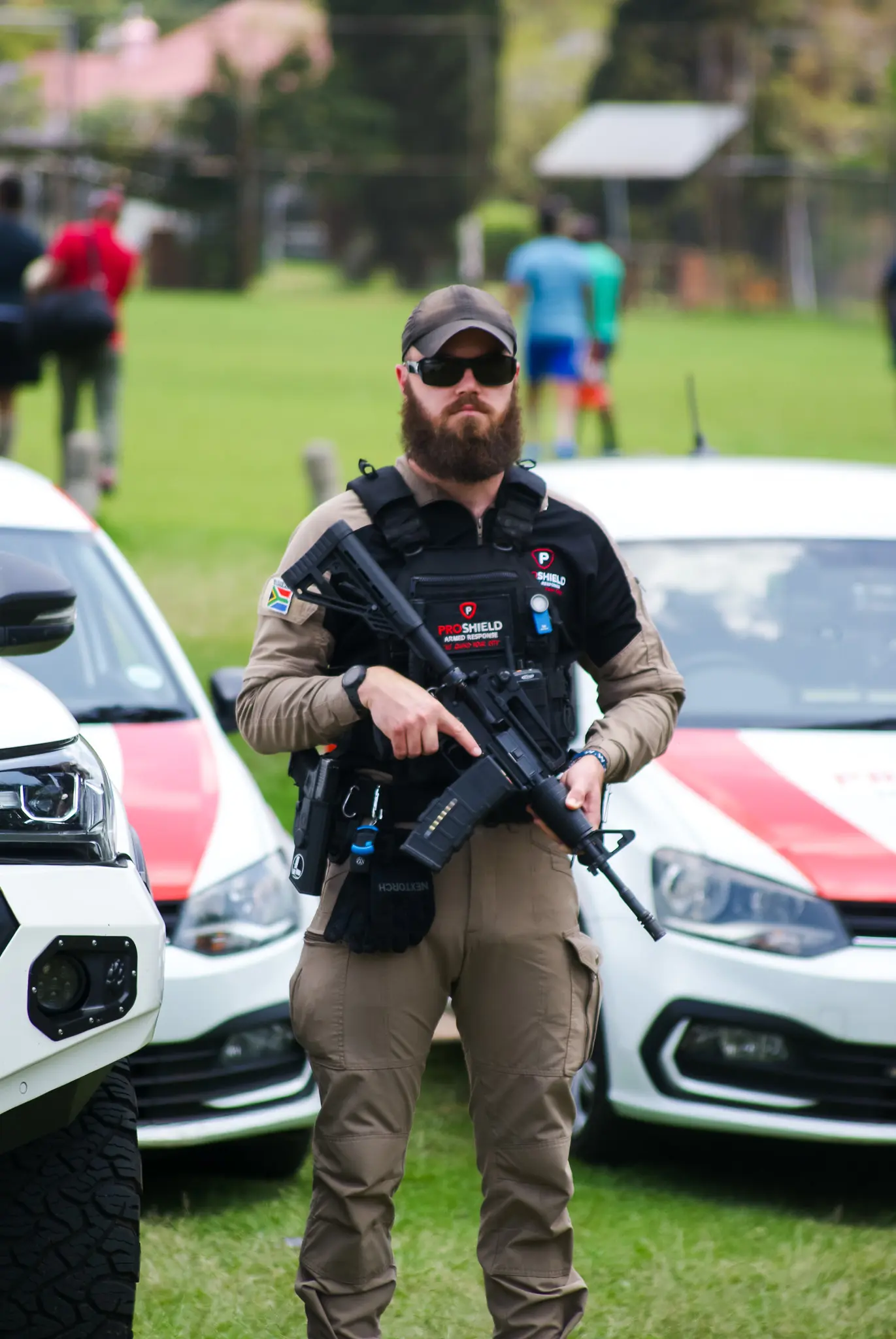 Proshield security guard standing with a gun infront of Proshield branded cars on a green grass field.